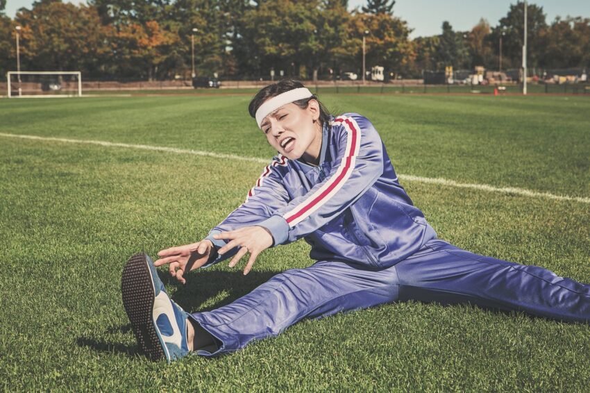 A woman trying to strech her leg in full fitness outfit, having pain to do so. Signifying that she is not fit for the task.